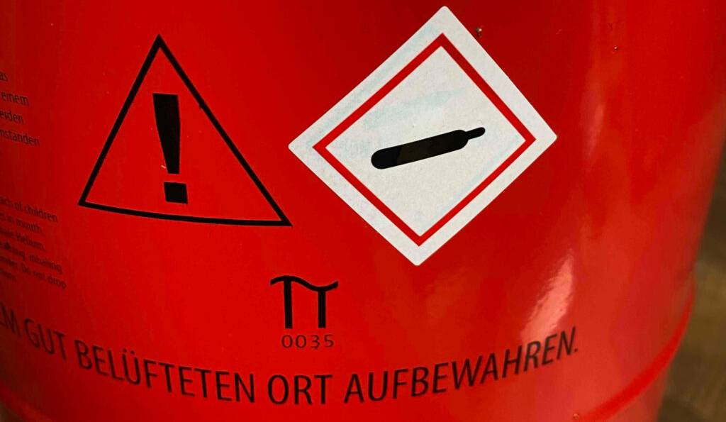 Ballongas-Test: Nahaufnahme eines roten Gasbehälters inklusive wichtiger Warnhinweise.