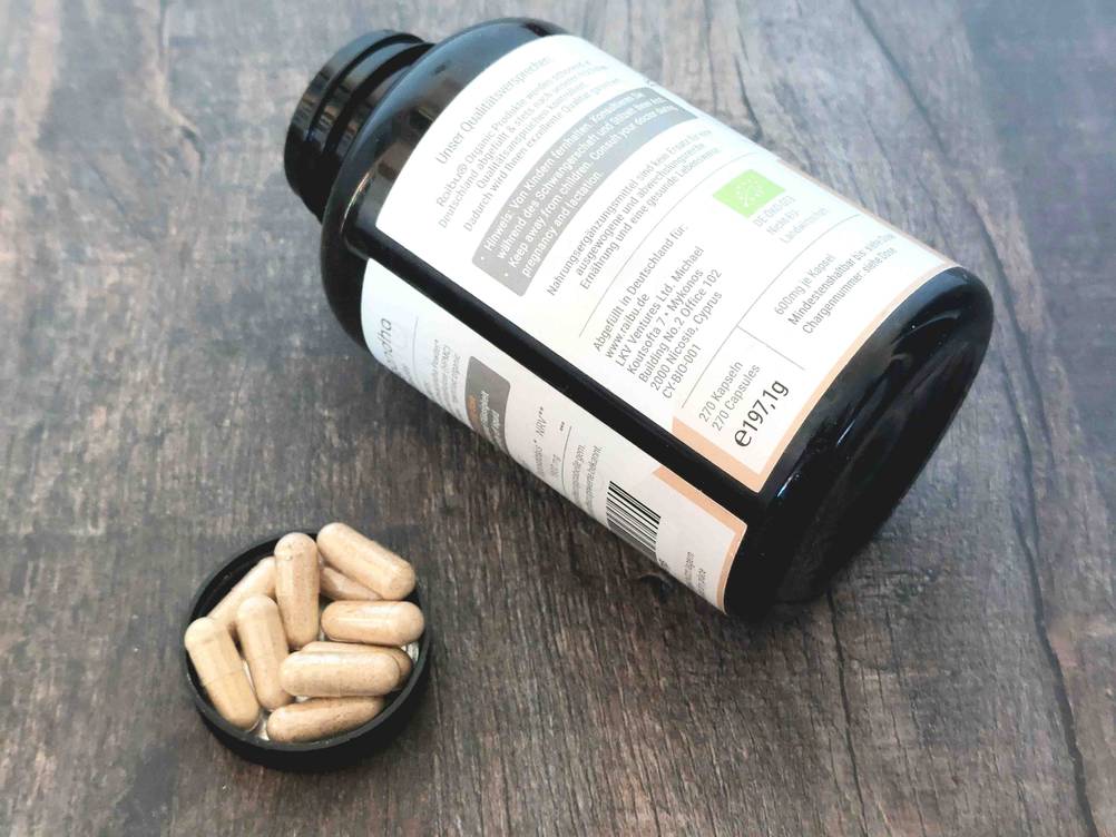 Rückseite einer Dose Ashwagandha mit Informationen auf einer dunklen Holzfläche, daneben einige Kapseln.