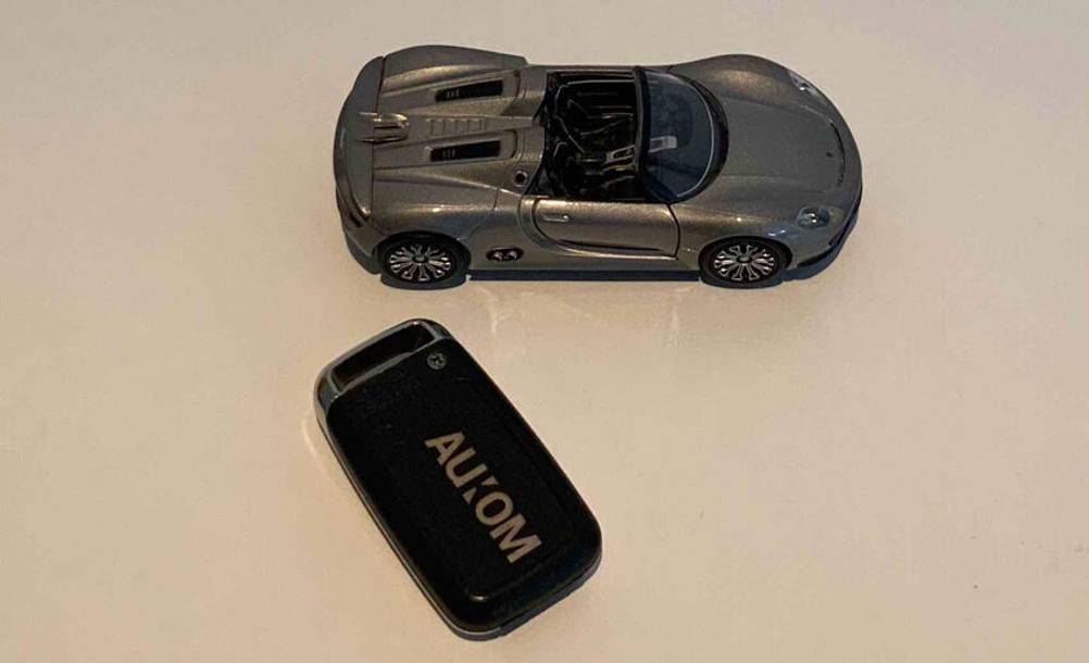 Ein schwarzer, getesteter Garagenöffner der Marke AUCOM liegt neben einem silberfarbenen Spielzeug-Rennauto auf einer weißen Oberfläche.