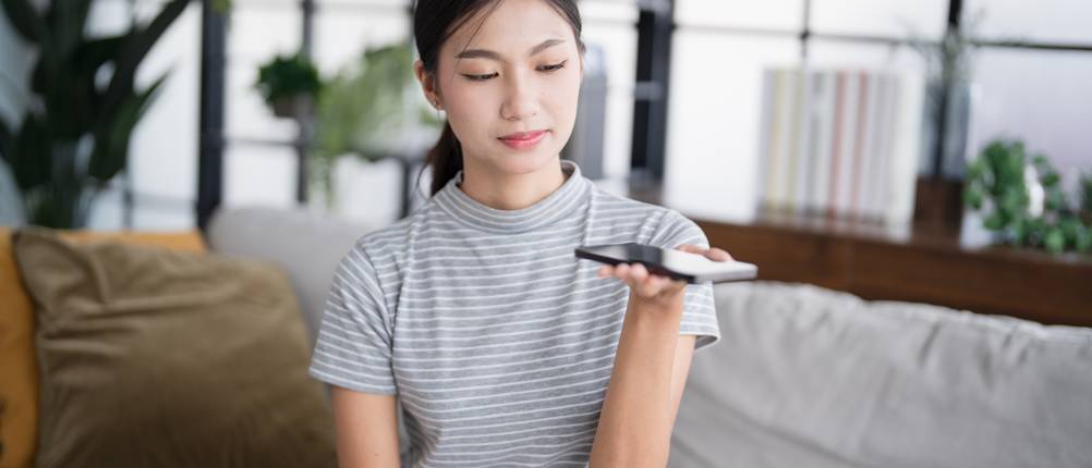 Audio-AI im Test: Eine Frau spricht in ein Smartphone, während eine Spracherkennungs-App aktiv ist. Im Hintergrund ein modernes Wohnzimmer mit Regalen.