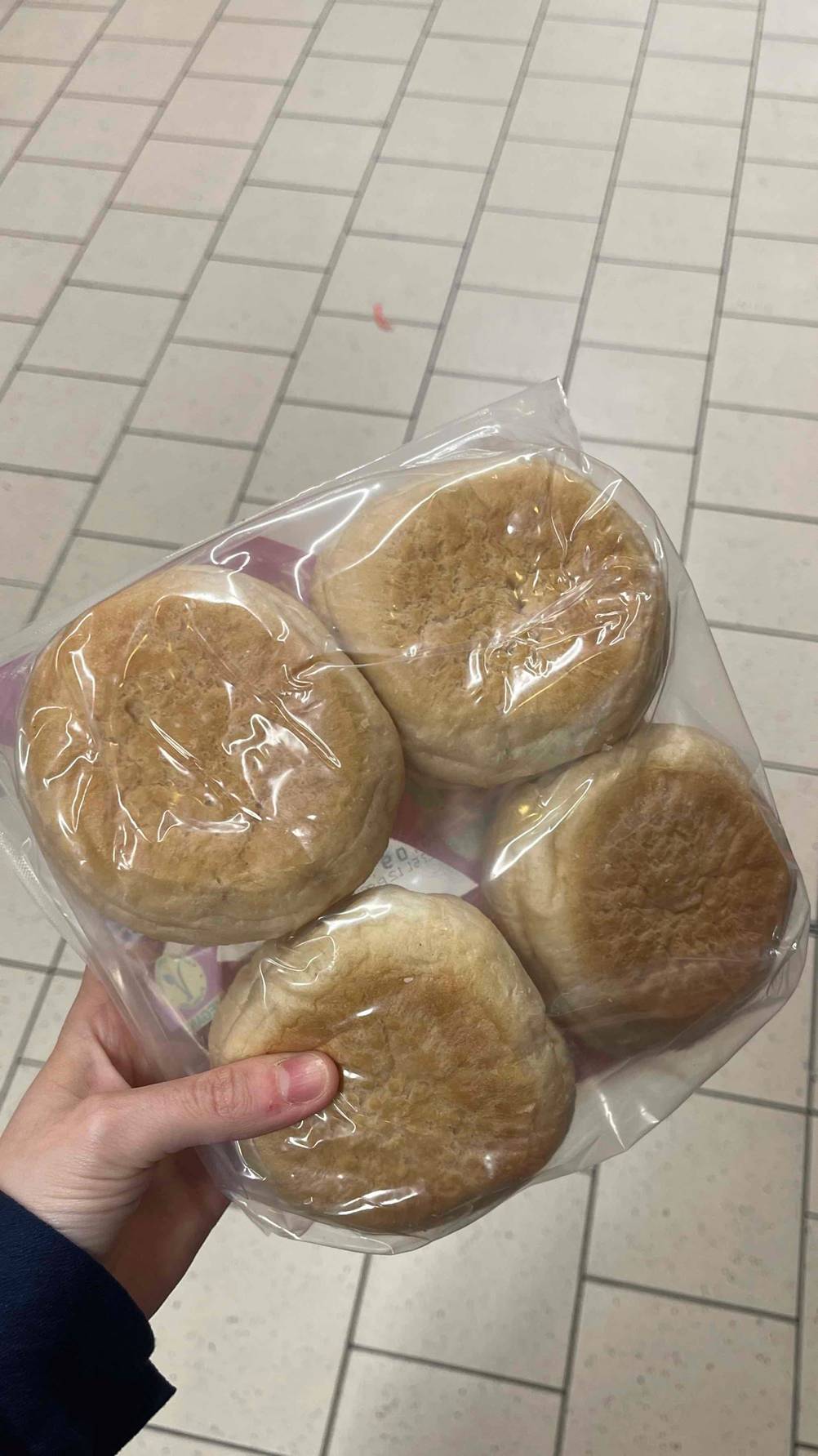 Eine Hand hält vier runde Aufbackbrötchen im Test innerhalb ihrer transparenten Kunststoffverpackung über einem weißen Fliesenbelag.