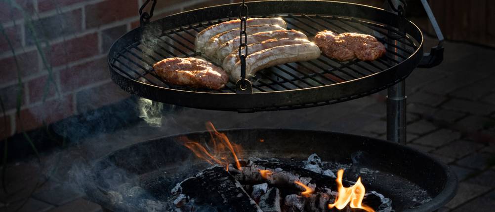 Ein Schwenkgrill mit Steaks und Würstchen steht vor einer Hauswand.