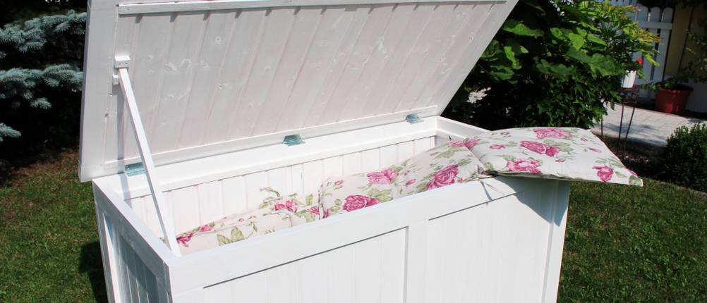 Geöffnete, weiße Aufbewahrungsbox für Sitzpolster steht in einem Garten. In ihr sind bereits Sitzpolster mit floralen Verzierungen verstaut.