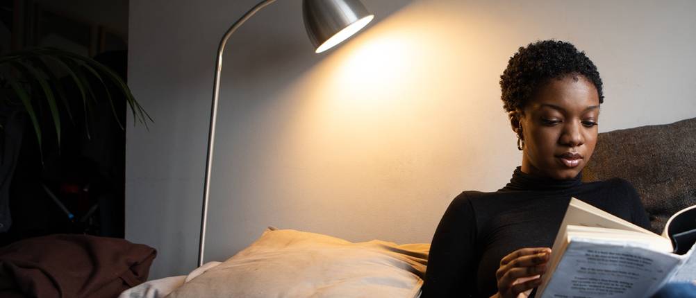 Frau liest auf Ihrer Couch in einem großen Buch, auf das eine Leselampe leuchtet.