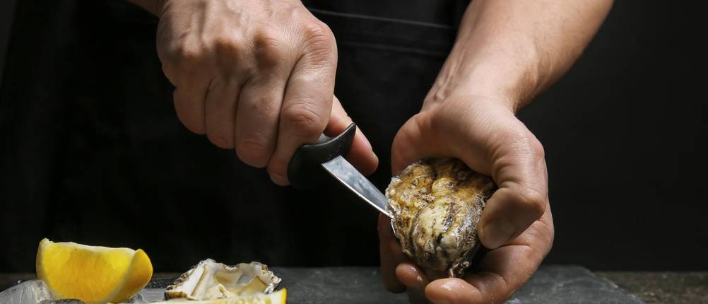 Austernmesser im Test: Mann öffnet Auster mit Austernmesser auf schwarzem Brett