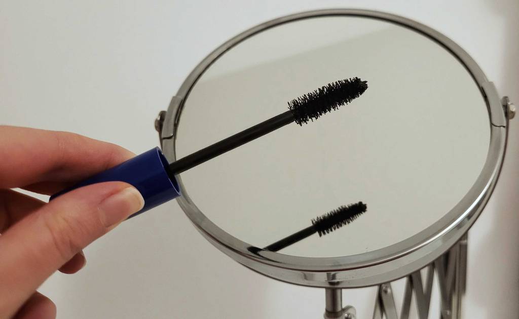 Hand hält eine offene Wimperntusche über der Vergrößerungsseite eines getesteten Kosmetikspiegels zur Wandmontage.