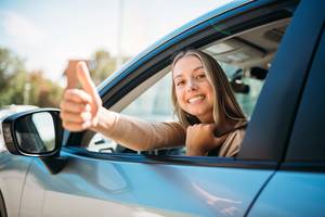 Mietwagenportale-Test: Eine junge Frau schaut aus dem Fenster eines Autos und streckt lächelnd den Daumen nach oben.