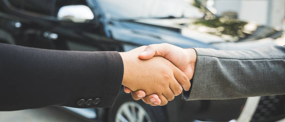 Seiten zum Auto verkaufen im Test: Zwei Personen schütteln sich die Hände, im Hintergrund ist ein Auto zu sehen.