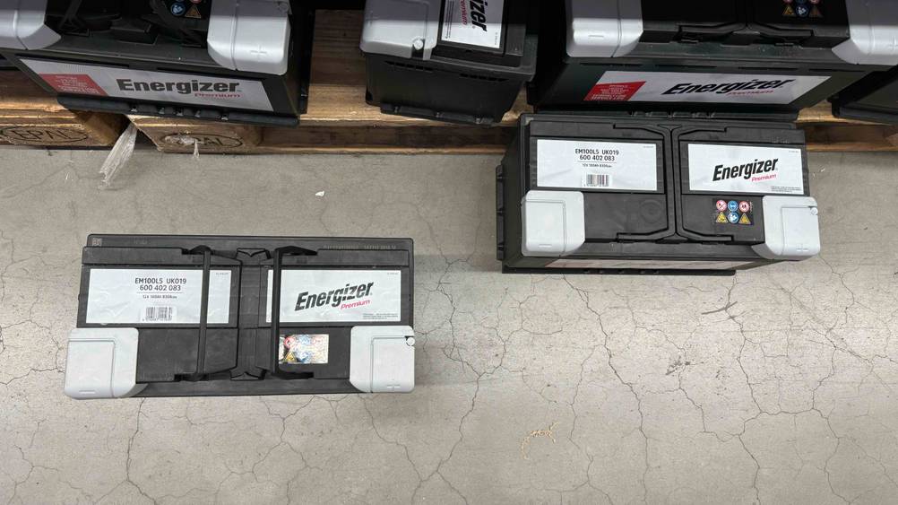 Zwei getestete Autobatterien 100Ah von oben auf einem Betonboden vor einem Regal stehend.