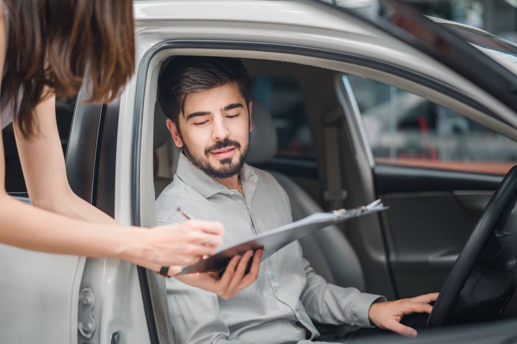 Autokredit im Vergleich: Ein junger Mann sitzt in einem Auto im Autohaus und blickt auf ein Angebot einer Verkaufsberaterin.