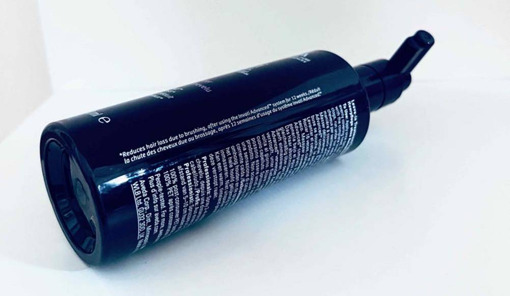 Eine schwarze Spender-Flasche mit Haarwuchsmittel im Test liegt mit Fokus auf die Anwendungsbeschreibung auf einer weißen Fläche.