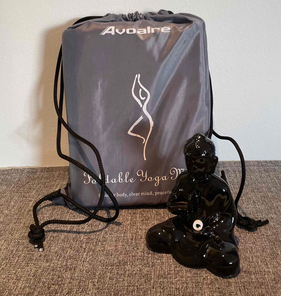 Getestet: Eine Reise-Yogamatte lehnt aufbewahrt im Beutel hinter einer schwarzen Buddha-Statue an einer weißen Wand.