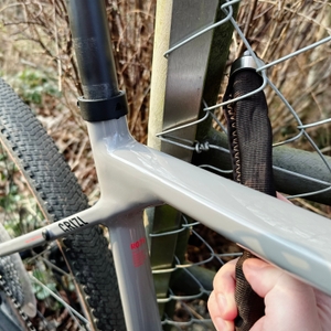 Das Axa Foldable 600 wird um den Fahrradrahmen und einen Zaun gelegt. Eine Hand zieht das Schloss durch das Gitter.