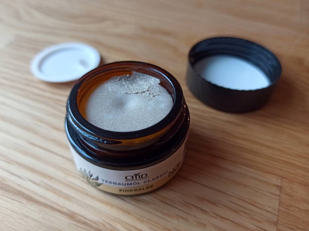 Eine geöffnete Anti-Pickel-Creme. Sie ist hellbräunlich. Daneben der Deckel und die entfernte Folie unter dem Deckel.