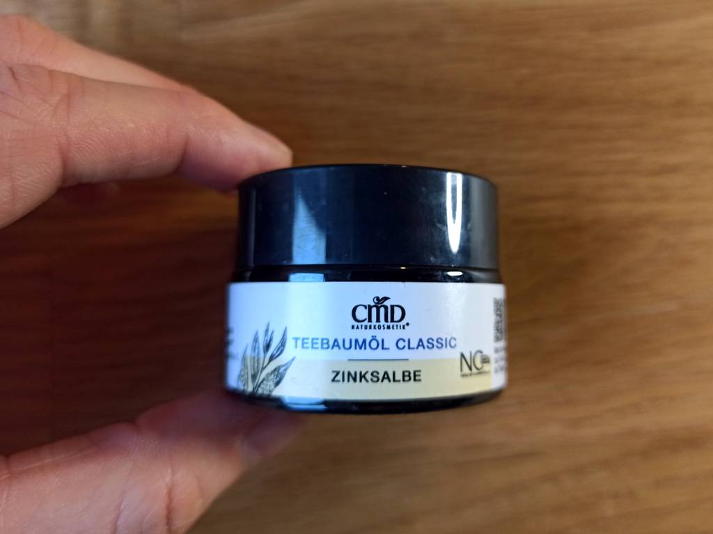 Eine Creme gegen Pickel in einer Hand, sie ist in einem dunklen Glastiegel verpackt und sehr klein.