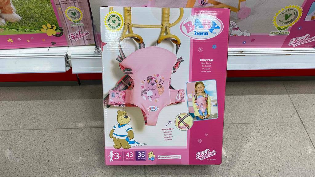 Getestet: Eine Verpackung mit einem Baby Born Puppentrage-Bild in Rosa steht auf einem grauen Boden vor einem Regal mit Babyprodukten.