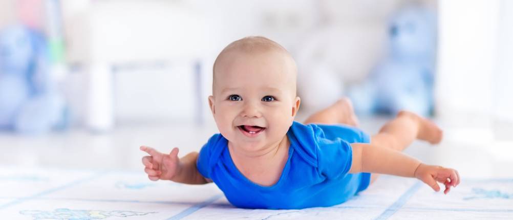 Adorable baby boy learning to crawl and playing with colorful toy in white sunny bedroom. Cute laughing child crawling on a play mat. Nursery interior, clothing and toys for little kids.