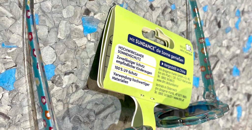 Detailansicht des Produktschilds an einer getesteten Baby-Sonnenbrille, die auf einer grauen Oberfläche liegt.