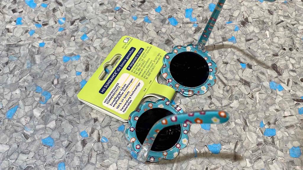 Baby-Sonnenbrillen-Test: eine Kinder-Sonnenbrille mit Kunststoff-Brillengestell in Hellblau, mit Blumen bedruckt, liegt auf einer grauen Oberfläche.