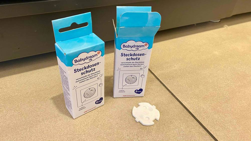 Eine Steckdosensicherung liegt zum testen vor einer geschlossenen und einer geöffneten Originalverpackung auf einem Fliesenboden.