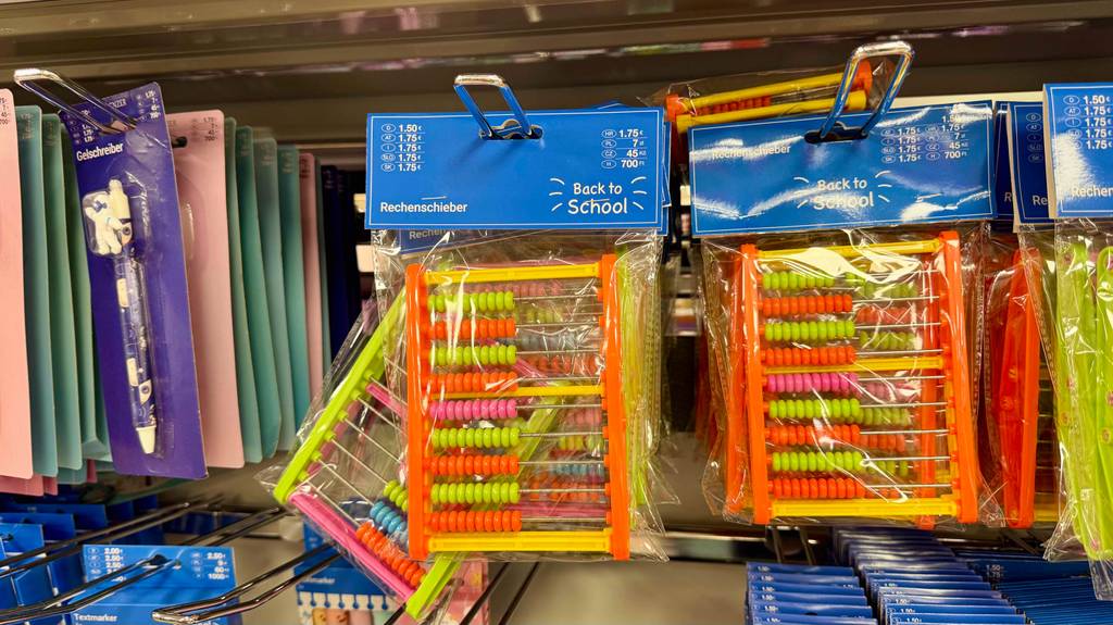 Mehrere verpackte Rechenschieber hängen zum testen in einem Regal zwischen anderen Schulartikeln.