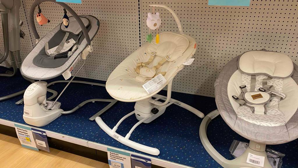 Vorderansicht einer elektrischen Babyschaukel im Test, die inklusive weißem Ständer und hellem Bezug mit Gurtsystem zusammen mit anderen Babyschaukeln im Verkaufsbereich steht.