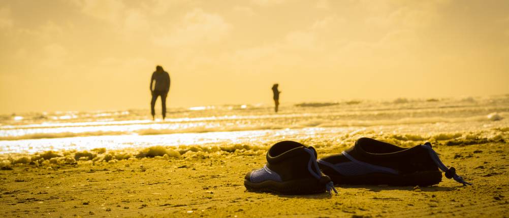 Badeschuhe für Erwachsene im Test: Strand-Szenerie in der Dämmerung, im Vordergrund 2 leere Badeschuhe, im Hintergrund 2 Personen im flachen Wasser