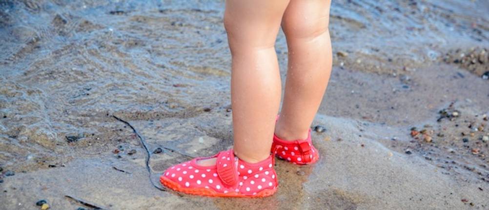 Kinder-Badeschuhe im Test: Beine von einem Kleinkind mit weiß gepunkteten roten Badeschuhen im Watt.
