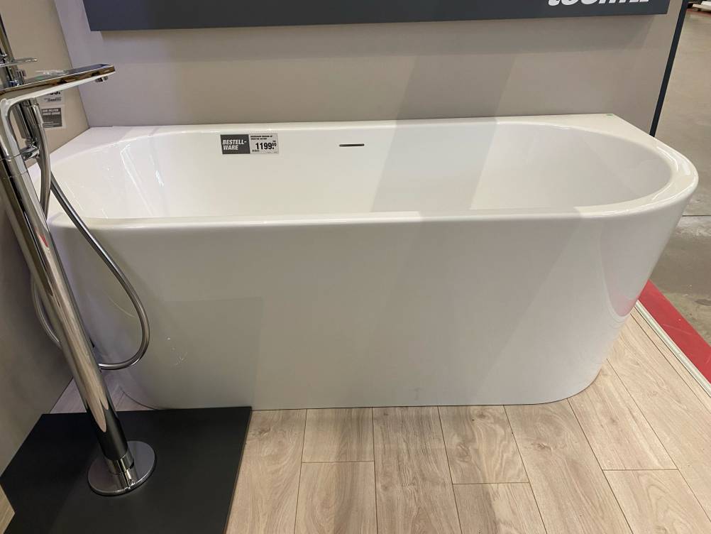 Ansicht der Längsseite einer weißen getesteten Duschbadewanne, die in einem Ausstellungsbereich an einer grauen Wand steht.