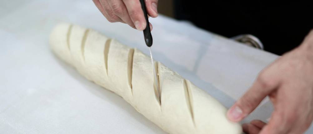 Bäckermesser im Text: Mann ritzt feine Schnitte in ein Baguette