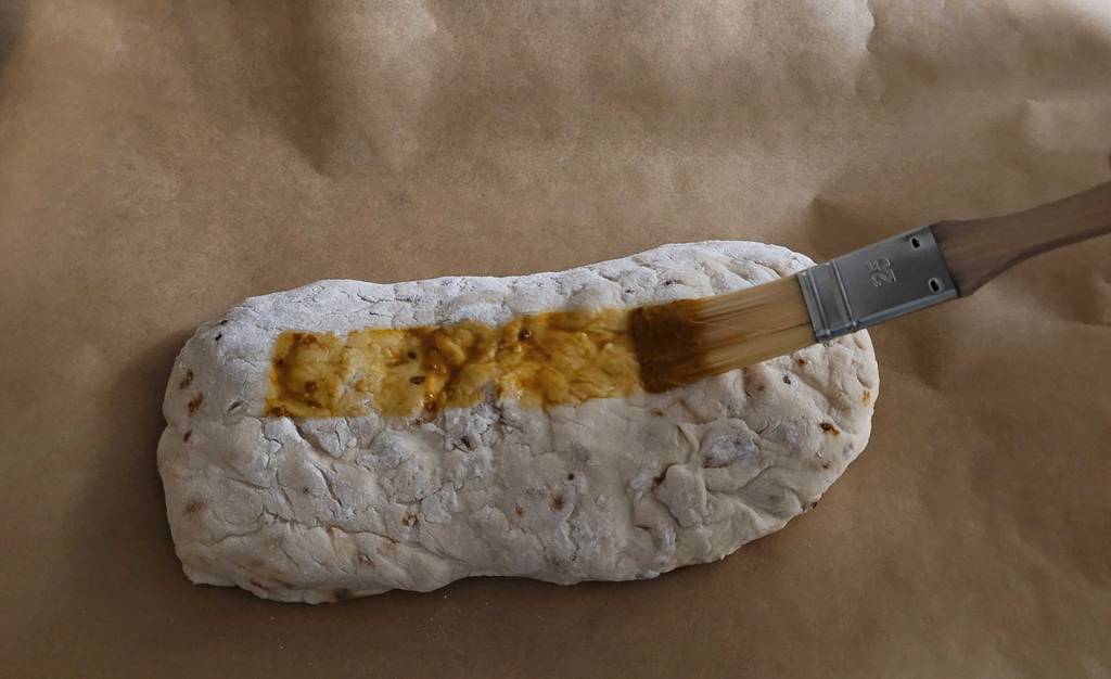 Dosenbrot im Test: Auf braunem Backpapier liegt ein zum Brot geformter Teig, der mit einem in Öl getränkten Pinsel bestrichen wird.