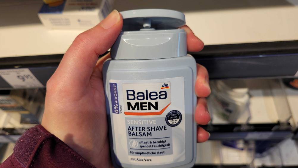 Hand öffnet den Klappverschluss einer hellblauen Flasche aus Kunststoff mit Balea Men Aftershave im Test, 0 % Alkohol, vor einem Verkaufsregal.