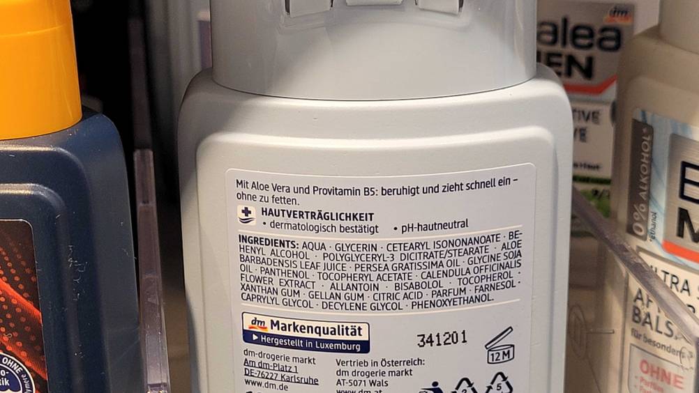 Detailansicht der rückwärtigen Produktbeschreibung einer hellgrauen Kunststoffflasche mit DM Aftershave im Test, in einem Verkaufsregal.
