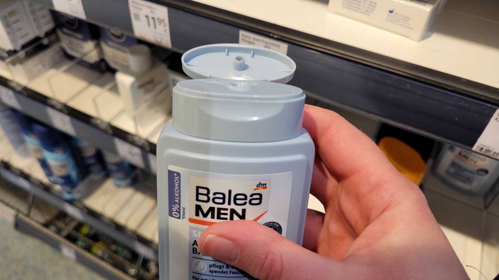 Hand hält eine hellblaue Kunststoffflasche, inklusive geöffnetem Klappverschluss, mit Balea Men Aftershave im Test, 0 % Alkohol, vor ein Verkaufsregal.