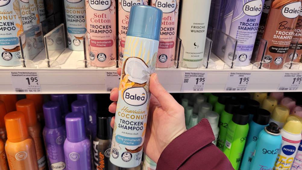 Trockenshampoo-Test: Eine Hand hält eine Dose vor ein Verkaufsregal mit anderen Produkten.