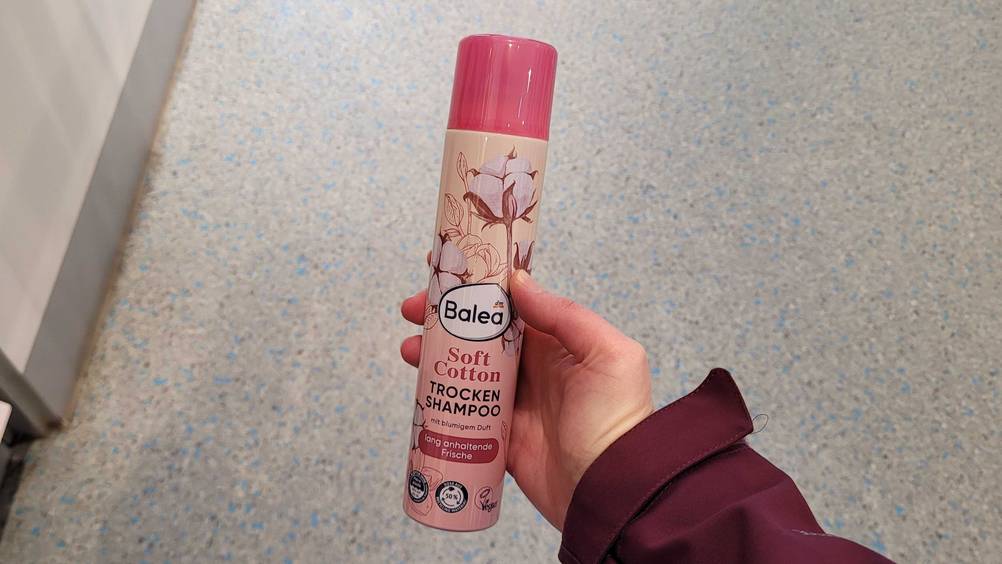 Eine Hand hält eine Dose Trockenshampoo im Test vor eine graue Fläche.