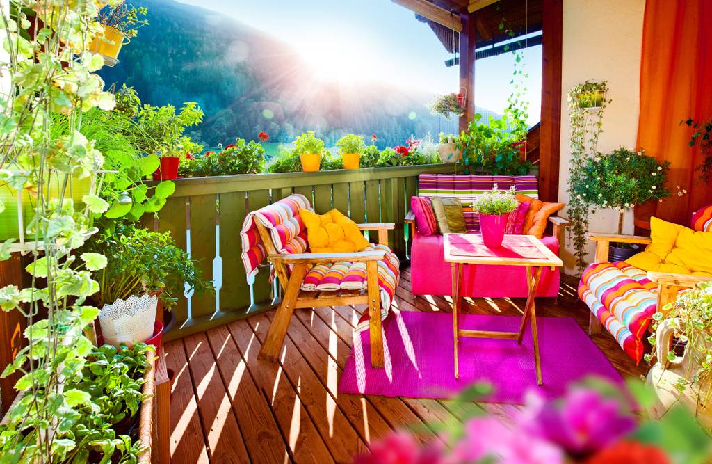 Auf dem Bild ist ein bunter Balkon mit vielen Pflanzen und bunten Kissen auf den Balkonmöbeln. Im Hintergrund ist ein Berg zu sehen.