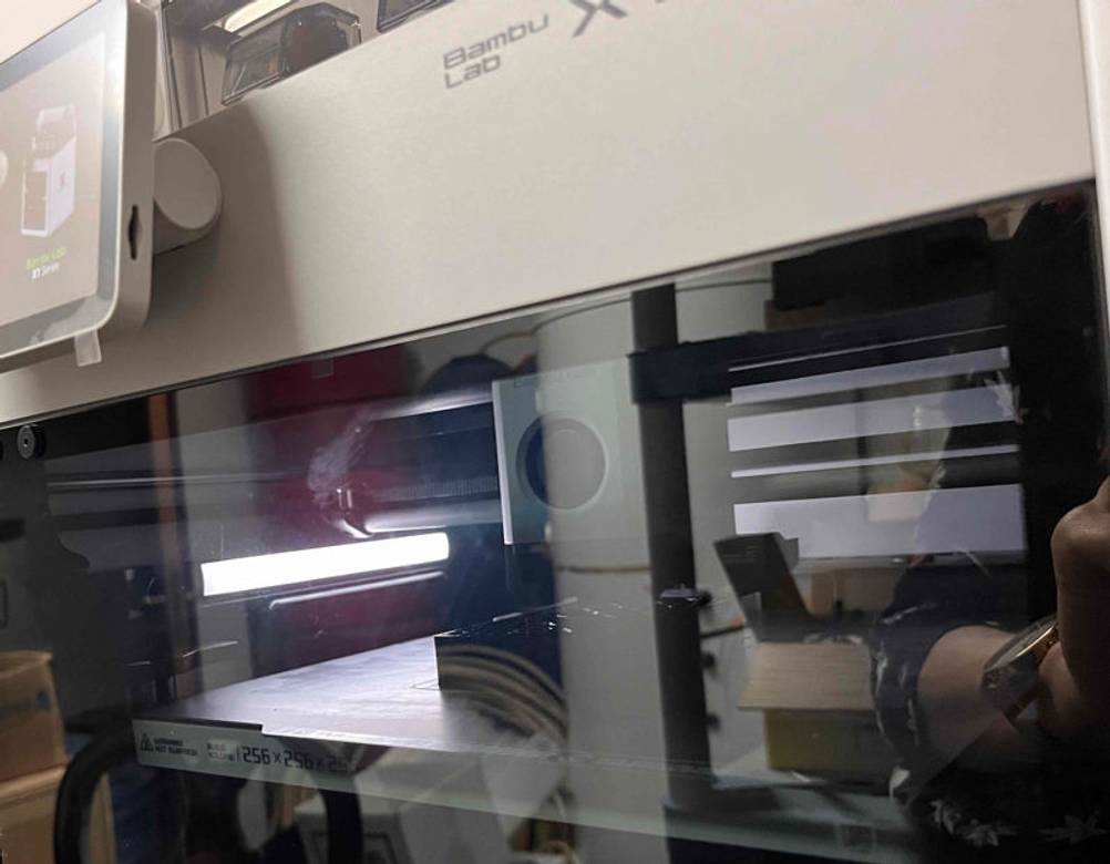 Nahaufnahme von einem Bambu Lab 3D-Drucker im Test, mit Innenbeleuchtung und einem auf Glas gespiegeltem Arm einer Person.