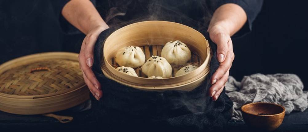 Bambusdampfer-Test: Zwei Hände umfassen einen Bambusdämpfer mit Dumplings. 