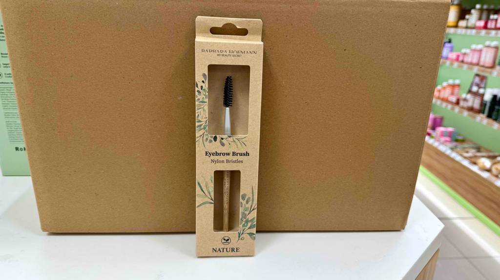 Ein Augenbrauenpinsel steht zum testen in Originalverpackung mit Sichtfenster vor einem Karton.