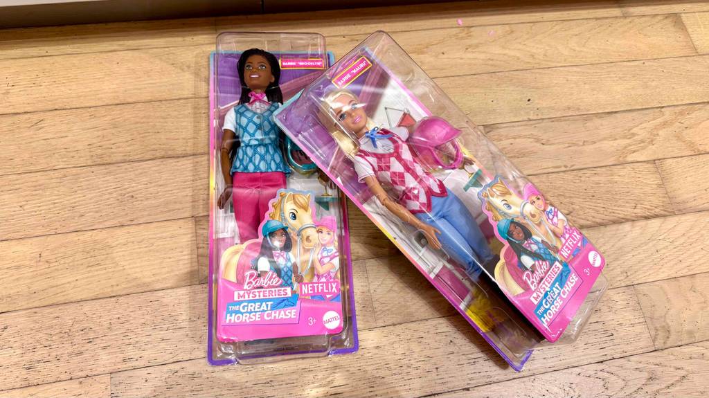 Zwei verschiedene Barbie-Puppen liegen in Originalverpackung zum testen auf einem Holzboden.