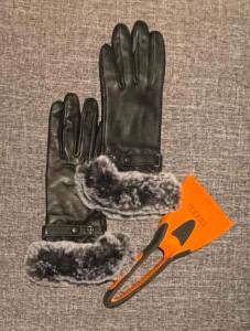 Schwarze Damen-Handschuhe aus Leder im Test mit grau meliertem, dekorativem Kunstfellbesatz liegen neben einem Eiskratzer auf einem anthrazitfarbenen Stoff.