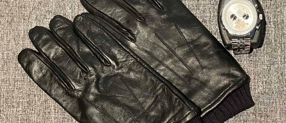 Schwarze getestete Herren-Handschuhe aus Leder mit elastischem Bund liegen neben einer eleganten Herrenuhr auf grauem Stoff.