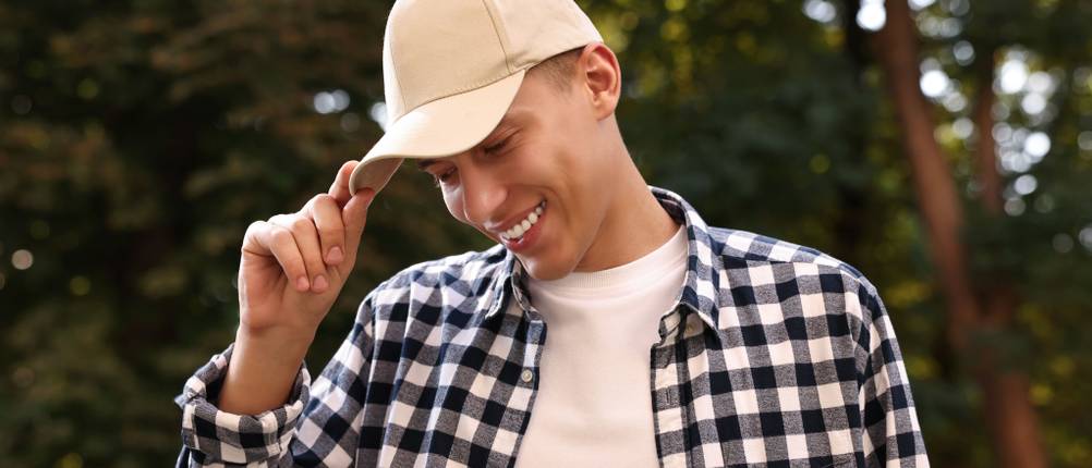 Baseball-Cap-Test: Ein junger Mann mit weißem T-Shirt und kariertem Hemd greift zum Schirm seiner weißen Baseball-Cap.