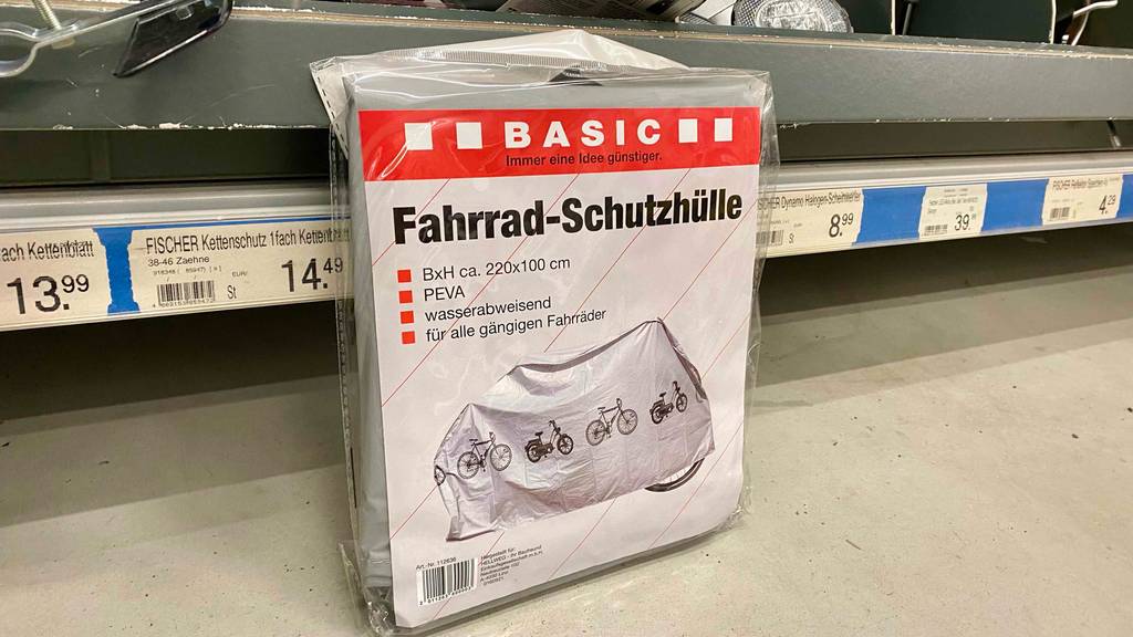 Ansicht einer grauen BASIC Fahrradschutzhülle im Test, die zusammengefaltet, mit schwarzen Zweirädern bedruckt, in einer Plastikverpackung auf dem Boden an einem Verkaufsregal lehnt.