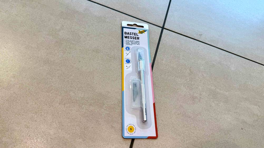 Auf einem Fließenboden liegt ein verpacktes Bastelmesser im Test.