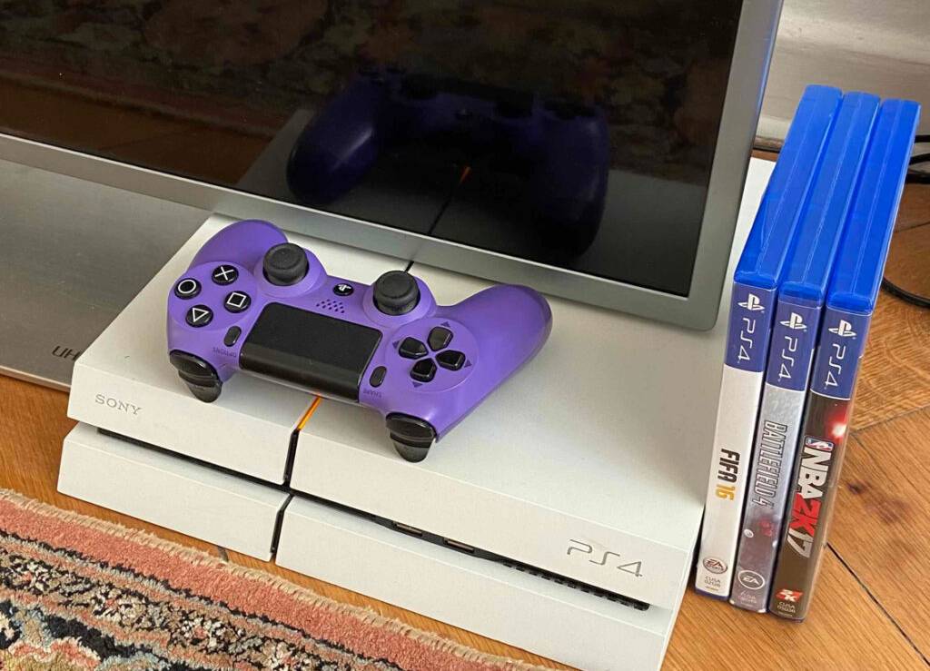 PS4-Spiele-Test: Mehrere Spiele in ihren Verpackungen stehen neben einem Monitor inklusive lilafarbenem PS4-Controller im Wohnbereich.