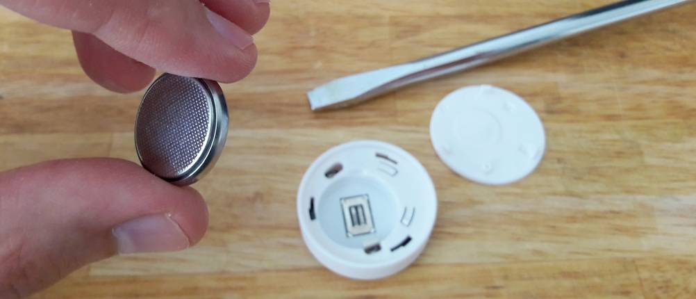 Eine Hand hält eine Knopfzelle, im Hintergrund geöffnetes Hygrometer