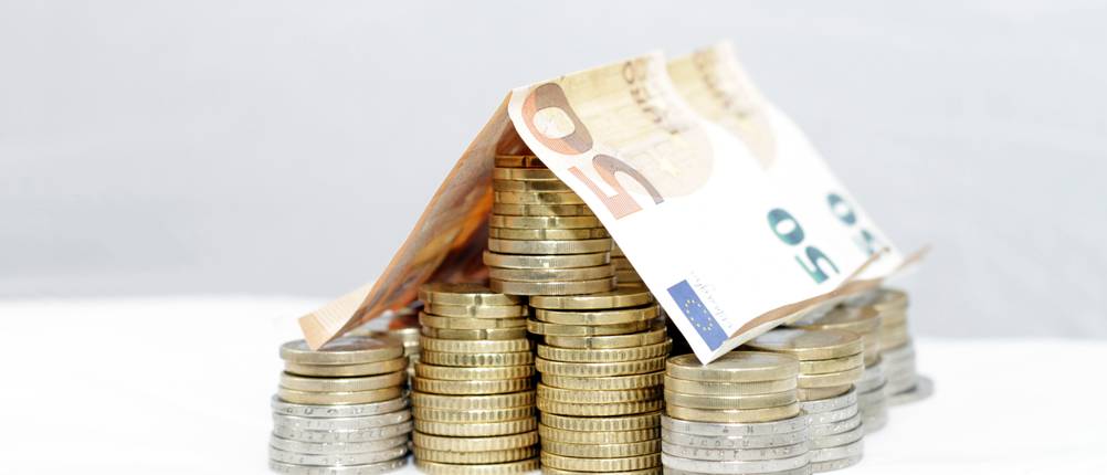 Auf einem Stapel Münzen liegen zwei 50-Euro-Scheine, die wie ein Dach geformt sind.