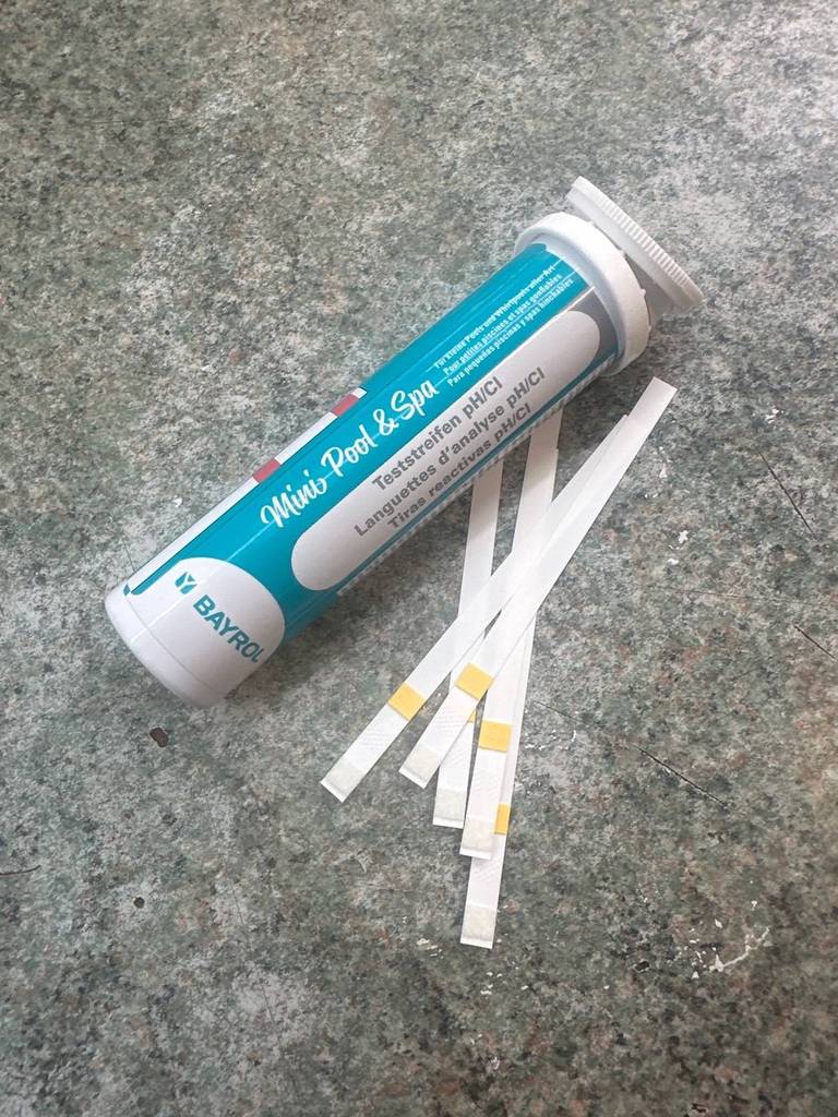 Pooltester im Test: Nahaufnahme einer türkis-weißen Dose der Marke Bayrol, neben der fünf Teststreifen auf grauem Boden liegen.